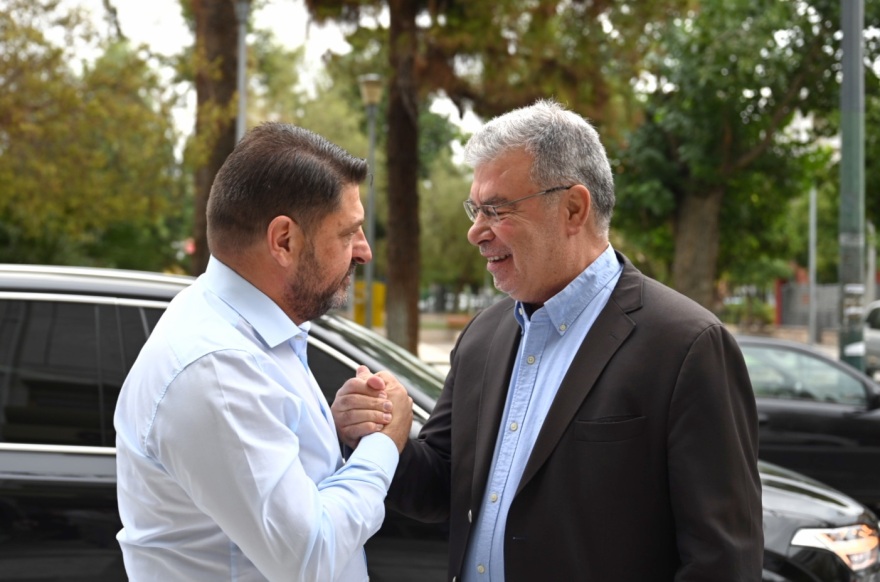 Δύο άνδρες χαιρετιούνται σε δημόσιο χώρο με φόντο δέντρα και αυτοκίνητα.